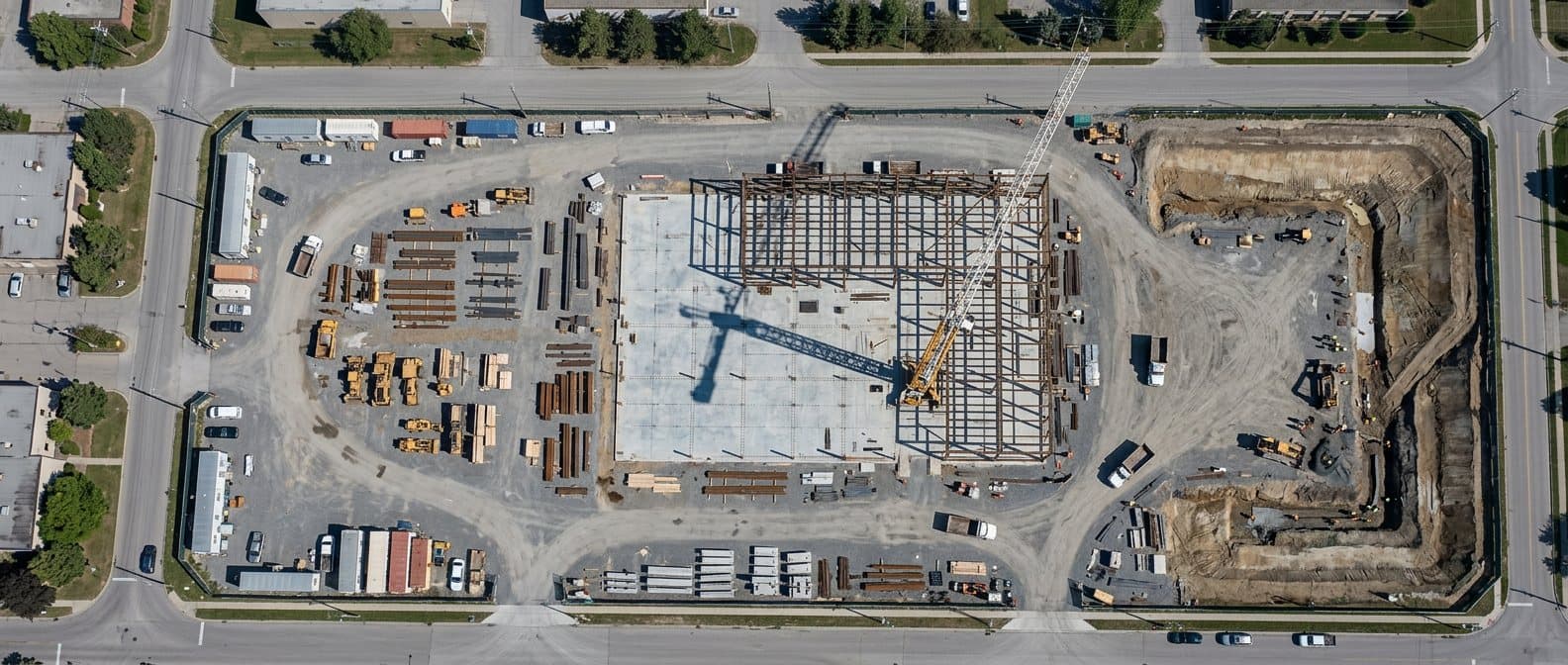 A construction site seen straight down and whole — slab, steel frame, laydown, excavation and haul roads in one continuous top-down view