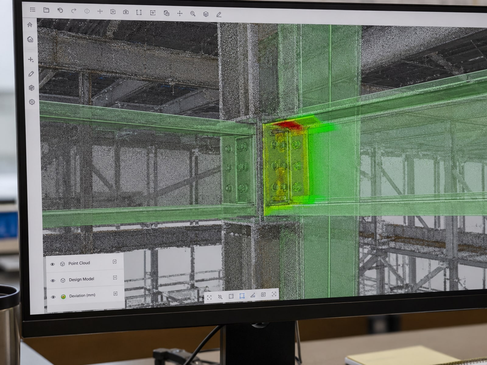 A monitor in a site office showing a grey point cloud of a steel frame with the design model overlaid in green, and a coloured patch across one beam-to-column connection where the two differ